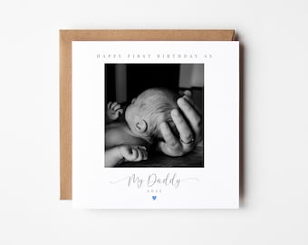 Puede incluir: Una foto en blanco y negro de un bebé acostado en las manos de una persona. La tarjeta dice "Feliz primer cumpleaños" y "Mi papá 2023" con un corazón azul.