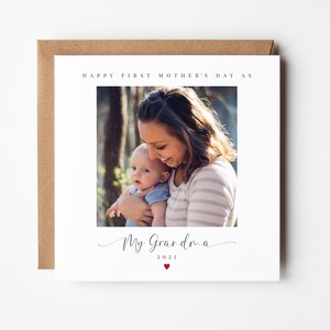 May include: A white card with a photo of a woman holding a baby. The text on the card reads "Happy First Mother's Day as My Grandma 2023" with a small red heart.
