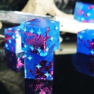 Könnte beinhalten: Eine Sammlung blauer Harzwürfel mit eingebetteten lila und silbernen Glitzer- und sternförmigen Akzenten. Die Würfel sind würfelförmig und haben eine reflektierende Oberfläche, mit einem dunklen Hintergrund.