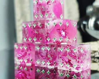 May include: A stack of six pink resin dice with white and green glitter embedded in the resin. The dice have a star pattern on each side.