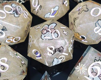 Op de afbeelding: Een set van zeven iriserende dobbelstenen met glitter ingebed in het heldere hars. De dobbelstenen zijn genummerd van 1 tot 6 en bevatten een verscheidenheid aan schattige cartoonafbeeldingen, waaronder een konijn, een strik en een bloem. De dobbelstenen zijn lichtbeige van kleur met witte nummers en afbeeldingen.