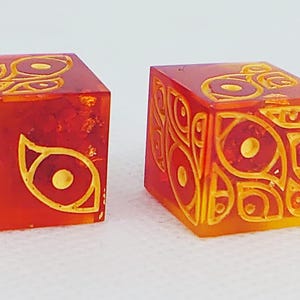 Op de afbeelding: Twee doorschijnende oranje en rode dobbelstenen met scherpe randen. Elke zijde van de dobbelstenen is voorzien van gegraveerde, geel ingevulde oogmotieven. De dobbelstenen hebben een glanzende afwerking en tonen een complex patroon van gestileerde ogen.