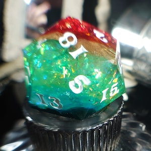 May include: A green, red, and gold iridescent dice with the numbers 13, 15, 8, 1, 5, and 3 on the faces. The dice is sitting on a black bottle stopper.