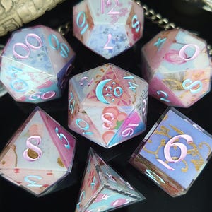May include: A set of seven polyhedral dice with a floral design. The dice feature a mix of pink, blue, and white colors with gold accents. The numbers are in a contrasting color, and the dice are arranged on a dark surface.