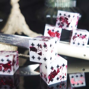 May include: White dice with red and black accents are stacked on a reflective surface. The dice have a star pattern and red inclusions. The dice are likely used for tabletop games.