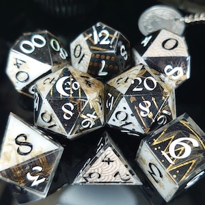 Op de afbeelding: Een verzameling polyhedrale dobbelstenen in verschillende vormen, waaronder d4, d6, d8, d10, d12 en d20. De dobbelstenen hebben een zwart-wit kleurenschema met gouden accenten en witte cijfers. Geschikt voor tabletop games.