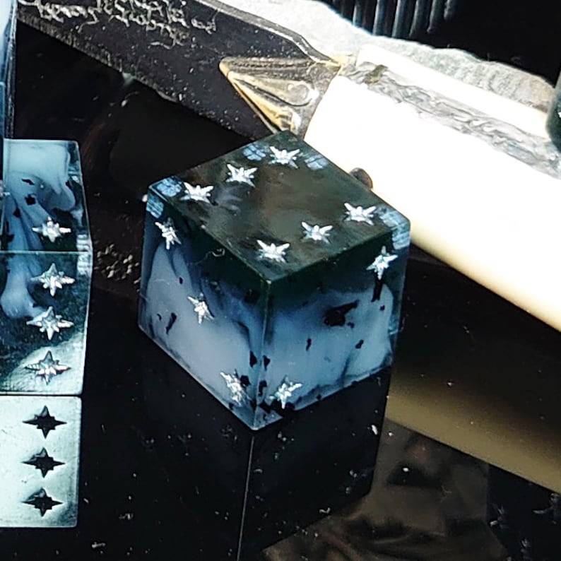 Pode incluir: Close-up de um dado azul escuro e verde com estrelas prateadas em cada lado. O dado &eacute; feito de resina e tem um efeito marmorizado.