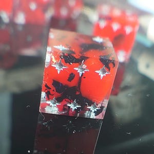 K&ouml;nnte beinhalten: Ein roter und schwarzer W&uuml;rfel mit silbernen Sternen auf jeder Seite. Der W&uuml;rfel ist aus klarem Harz gefertigt und hat einen Marmoreffekt.