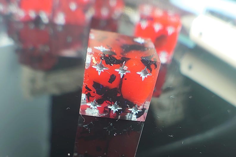 K&ouml;nnte beinhalten: Ein roter und schwarzer W&uuml;rfel mit silbernen Sternen auf jeder Seite. Der W&uuml;rfel ist aus klarem Harz gefertigt und hat einen Marmoreffekt.