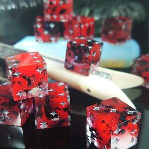 K&ouml;nnte beinhalten: Ein Satz roter W&uuml;rfel mit schwarzen und silbernen Akzenten. Die W&uuml;rfel sind aus klarem Harz gefertigt und haben ein wirbelndes Muster im Inneren. Die W&uuml;rfel sind auf einer schwarzen Oberfl&auml;che verstreut.