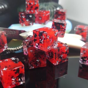 K&ouml;nnte beinhalten: Eine Gruppe roter W&uuml;rfel mit schwarzen und silbernen Flecken. Die W&uuml;rfel sind aus Harz und haben eine klare Oberfl&auml;che. Sie sind auf einer schwarzen Oberfl&auml;che verstreut.