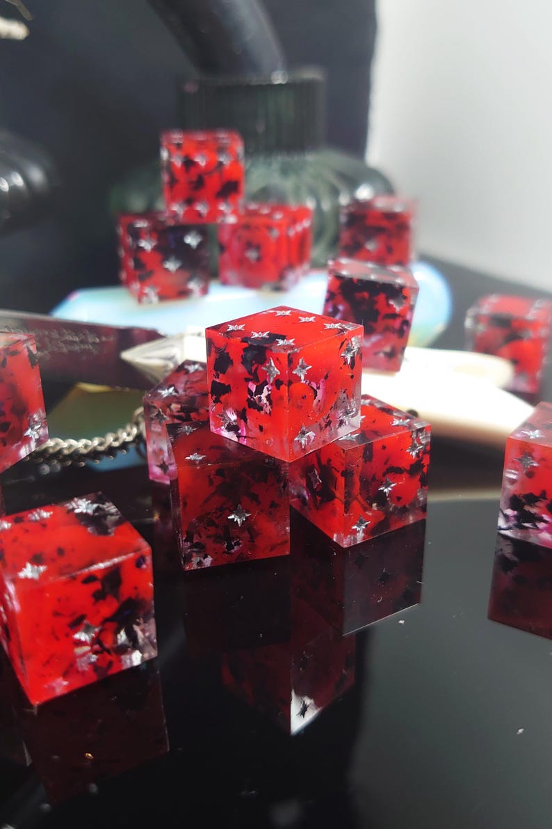 K&ouml;nnte beinhalten: Eine Gruppe roter W&uuml;rfel mit schwarzen und silbernen Flecken. Die W&uuml;rfel sind aus Harz und haben eine klare Oberfl&auml;che. Sie sind auf einer schwarzen Oberfl&auml;che verstreut.