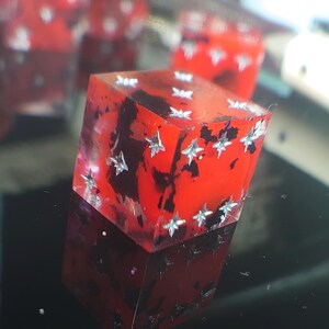 K&ouml;nnte beinhalten: Ein roter W&uuml;rfel mit silbernen Glitzersternen, die in das Harz eingebettet sind. Der W&uuml;rfel ist durchscheinend und hat schwarze Wirbel im Inneren.