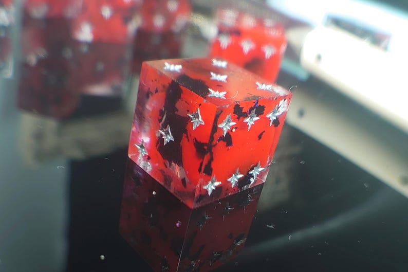 K&ouml;nnte beinhalten: Ein roter W&uuml;rfel mit silbernen Glitzersternen, die in das Harz eingebettet sind. Der W&uuml;rfel ist durchscheinend und hat schwarze Wirbel im Inneren.