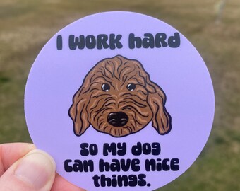 May include: A round sticker with a light purple background and the text "I WORK hard SO MY dog CAN HAVE nice things." A brown doodle dog illustration is in the center. The sticker is held by a hand.