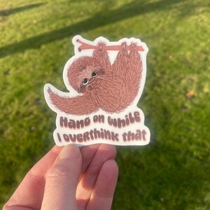 Puede incluir: Una pegatina de perezoso marrón con el texto "Hang on while I overthink that" en una fuente retro.