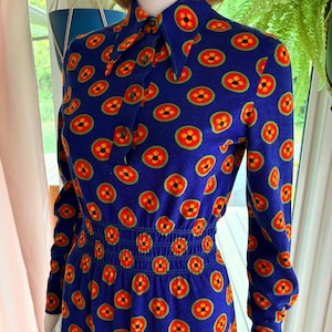 May include: A long-sleeved dress in a deep blue fabric featuring a repeating pattern of circular motifs. Each motif consists of a bright orange outer ring, a green border, and a yellow four-petaled flower center. The dress has a pointed collar, buttons.