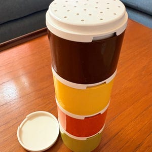 May include: A colorful, vintage food storage container with a white lid and a stack of four sections in brown, yellow, red, and green. The lid has small holes. A separate white lid is next to the container.