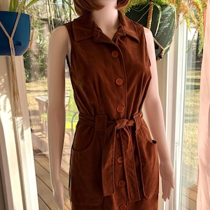 May include: A sleeveless, button-down dress in a rich brown hue. The dress features a collared neckline, a belted waist, and two front pockets. The dress is displayed on a mannequin, showcasing its vintage style. The mannequin is wearing a brown wig.