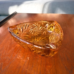 May include: Amber-coloured glass ashtray with a triangular shape. The ashtray features intricate floral and geometric patterns etched into the glass. The ashtray is sitting on a wooden surface.