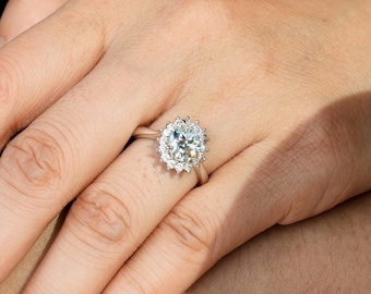 Peut inclure: Une bague en argent avec une grande pierre précieuse ronde et transparente entourée de petites pierres précieuses transparentes. La bague est portée au doigt d'une personne.