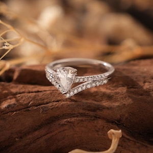 May include: A pear-shaped diamond engagement ring set in a silver band with a V-shaped design. The ring is set on a piece of brown driftwood.