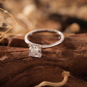 Peut inclure: Une bague en argent avec un diamant de forme carrée serti au centre. La bague repose sur un morceau de bois brun.