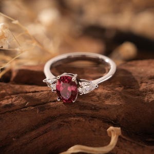 May include: A silver ring with a large oval-shaped red gemstone and two smaller clear gemstones on either side.