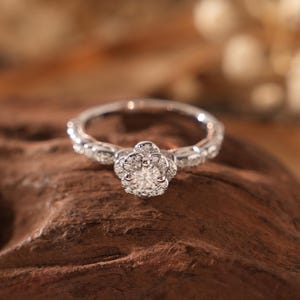 May include: A silver engagement ring with a floral design. The ring features a central, round-cut diamond surrounded by smaller stones, set on a band with intricate detailing. The ring is displayed on a brown wooden surface.