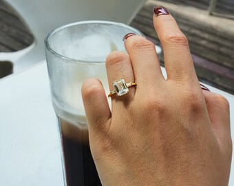 May include: A close-up of a hand wearing a gold ring with a large, clear emerald-cut diamond. The ring is on the index finger of the hand. The hand is resting on a glass of iced coffee.