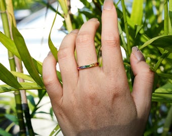 May include: A gold-colored ring with a row of colorful gemstones, including red, orange, yellow, green, blue, and purple. The ring is set against a backdrop of green bamboo stalks and leaves.