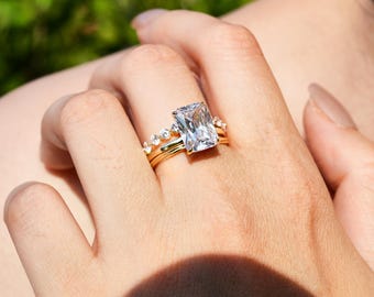 May include: A gold ring with a large, clear, rectangular-shaped gemstone. The ring is set with smaller gemstones around the band.