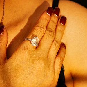 May include: A close-up of a hand wearing a silver ring with a large oval-shaped diamond. The ring has a halo of smaller diamonds around the center stone.