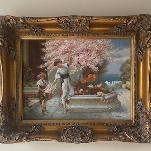 Puede incluir: Pintura enmarcada con un marco dorado ornamentado, que representa a dos figuras recogiendo flores bajo un árbol de flores rosadas. La obra presenta una escena de jardín clásico con una fuente y un edificio distante, un tema sereno y pastoral.