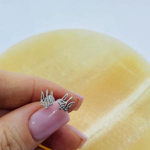 May include: A pair of silver-toned stud earrings featuring the Ukrainian Tryzub emblem. The earrings have a polished metallic finish and a cutout design that forms the shape of the trident symbol, each secured with a small circular backing.