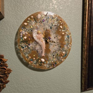 May include: A round, brown resin clock with a white angel figurine and gold accents. The clock face is decorated with seashells, glitter, and other embellishments.