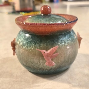 May include: A decorative ceramic jar with a lid. The jar is teal and tan with pink hummingbird accents. The lid is orange with a small, round, orange knob. The jar is sitting on a light-colored surface.