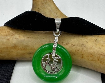 May include: A green jade pendant with a silver charm featuring Chinese characters. The pendant is on a black velvet cord.