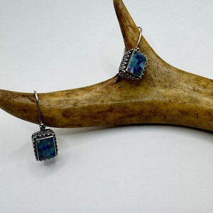 May include: Silver-toned earrings with square, mosaic-style blue and green iridescent stones. The earrings are suspended from a simple hook and are displayed on a light brown antler against a white background. Jewelry.