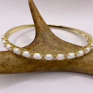 May include: A gold bangle bracelet featuring a row of white opal stones set in a gold setting. The bracelet is displayed on a brown antler.