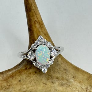 Könnte beinhalten: Ein silberner Ring mit einem weißen Opal-Edelstein, der von kleineren, klaren Edelsteinen umgeben ist.