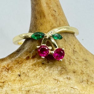 May include: A gold ring with a cherry motif. Two round, bright pink gemstones form the cherries, with green marquise-cut gemstones as leaves. The ring is displayed against a neutral background.