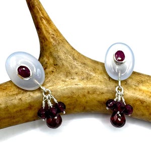 May include: A pair of silver earrings featuring a white stone and a red gemstone. The earrings have a cluster of small red gemstones dangling from the bottom.