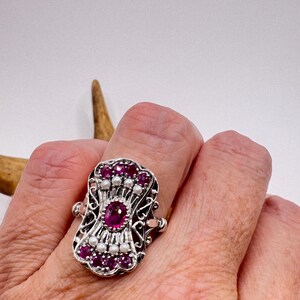 May include: A silver ring featuring a central oval ruby, surrounded by smaller rubies and pearls. The ring has an elaborate, filigree design. The ring's face is rectangular.
