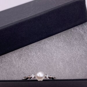 May include: A silver ring with a white pearl and small clear stones set in a delicate design. The ring is displayed in a black gift box with white padding.