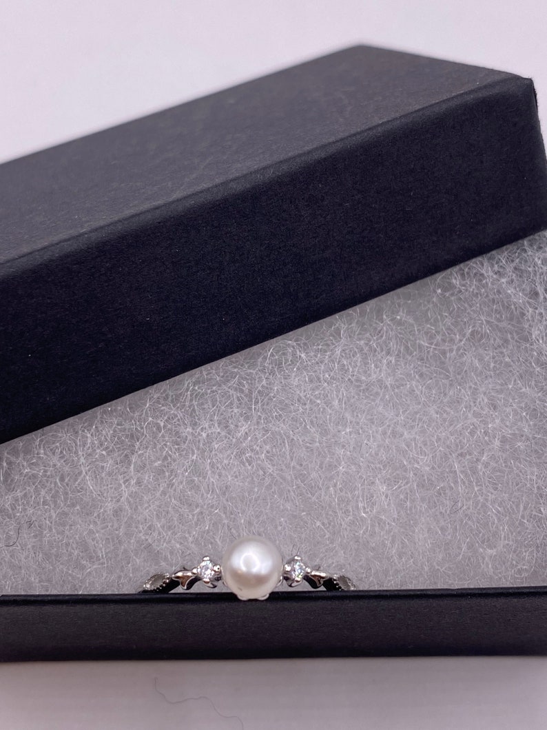 May include: A silver ring with a white pearl and small clear stones set in a delicate design. The ring is displayed in a black gift box with white padding.