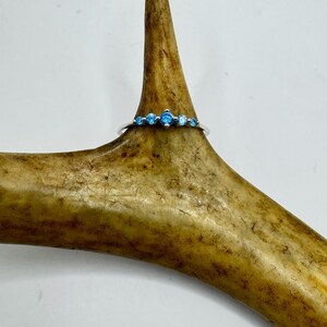 May include: A silver ring with five light blue gemstones. The ring is displayed on a brown antler against a white background. The gemstones are evenly spaced across the top of the ring.