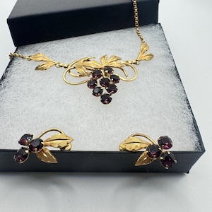 May include: A gold-tone metal jewellery set consisting of a necklace and a pair of screw-back earrings. The necklace features a grape cluster motif with purple faceted stones and textured leaf accents. The earrings mirror the grape and leaf design.