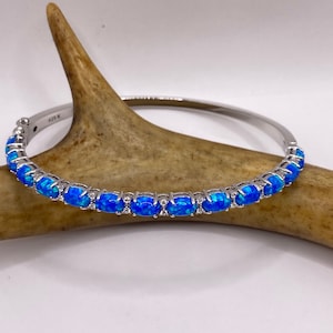 May include: A silver bangle bracelet with a row of blue oval-shaped opals set in a silver band. The bracelet is on a brown antler.