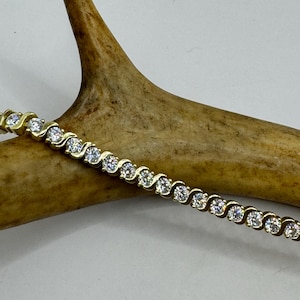 Op de afbeelding: Een gouden tennisarmband met een golvend ontwerp en heldere ronde stenen.